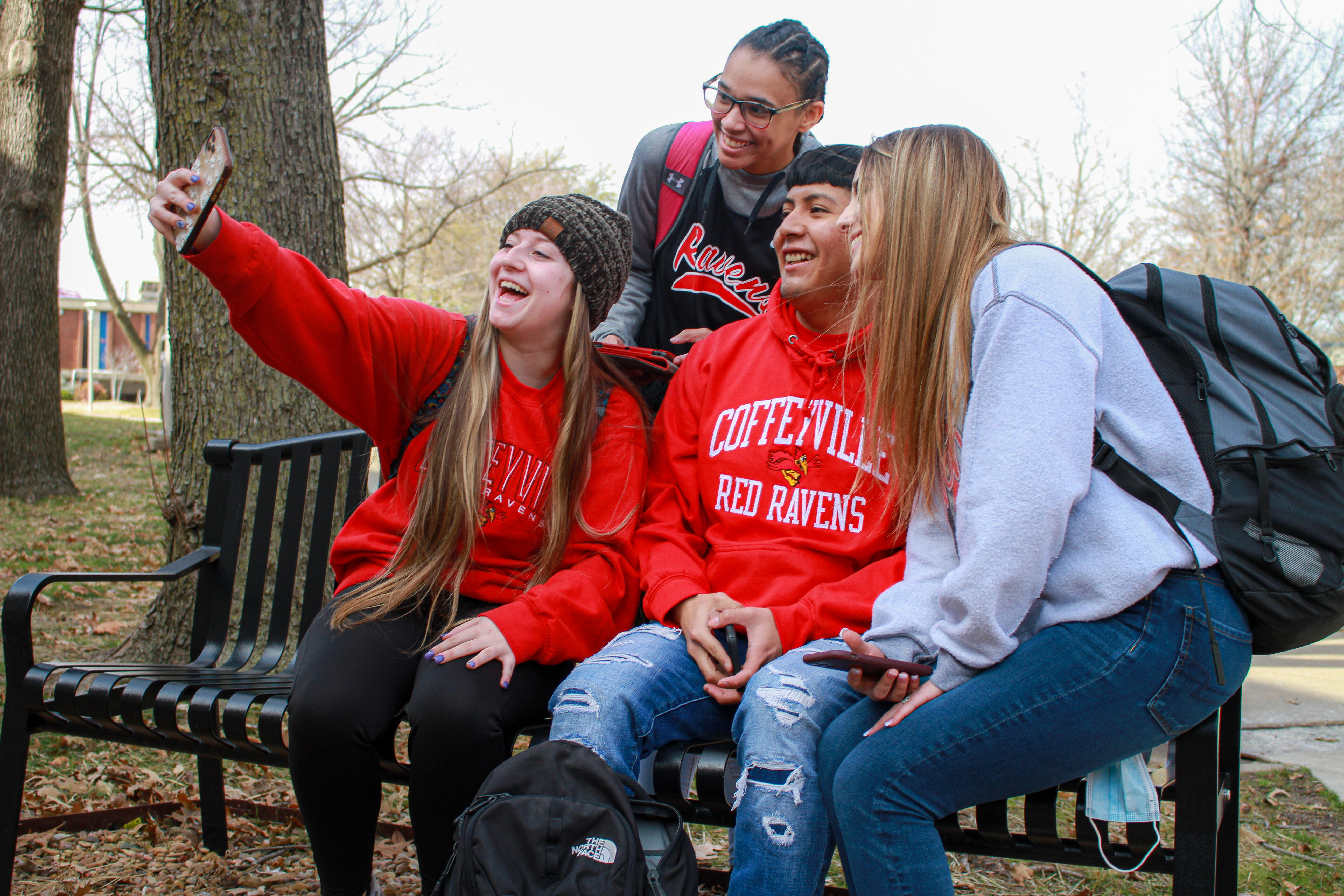Four students sitting on a bench in the square taking a selfie